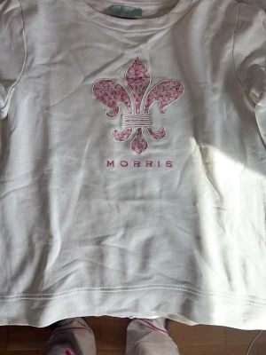 Vit t-shirt från Morris med blommigt tryck - Vit t-shirt från Morris med ett stort blommigt tryck i rosa på bröstet och broderad detalj på ärmen. Klassisk rund hals och korta ärmar. Perfekt för dig som gillar stilrena plagg med en twist. Materialet är mjuk bomull.Äkta!