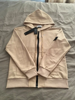 Beige hoodie med dragkedja från stone island - Säljer en beige hoodie från Stone island med dragkedja framtill och två fickor. Hoodien har ribbade muddar och är tillverkad i mjukt bomullsmaterial. Perfekt för en avslappnad och stilren look. Huva med snörning och diskret logotyp på ärmen.