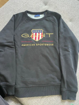 Svart sweatshirt från GANT - Cool svart sweatshirt från GANT med broderad logga och emblem på bröstet. Tröjan har rund halsringning, ribbade muddar och texten 'American Sportswear' i gult. Perfekt för en avslappnad och sportig look. 