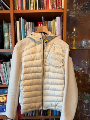 Parajumper cardigan  - Snygg beige pufferjacka från Parajumpers med huva och dragkedja framtill. Jackan har quiltad front och rygg, samt stickade ärmar och sidopartier. Bröstficka med dragkedja och klassisk Parajumpers-logga på ärmen. Perfekt för kalla vinterdagar, vårdagar och höstdagar. (Alla taggar finns kvar och kvittot finns den är köpt på NK)