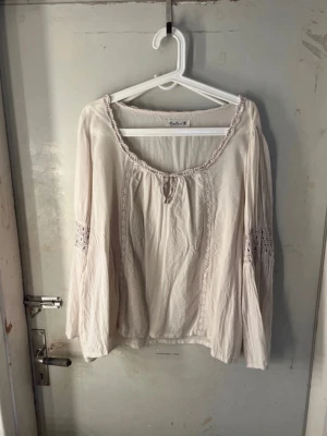 Blus  - Söt krämvit blus  med långa, vida ärmar och fina spetsdetaljer längs halsringningen och på ärmarna. Blusen har en avslappnad passform och rundad hals med liten knytning framtill. Perfekt för en avslappnad och trendig look.
