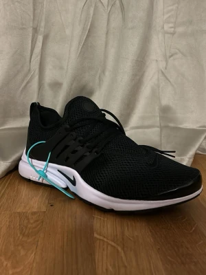 Nike Air Prestos - Svart sneaker från Nike med mesh-överdel och vita detaljer på sulan. Skon har klassiska snörningar och en låg profil. Sidan pryds av Nike-loggan och plastdetaljer vid snörningen. Perfekt för dig som gillar sportig och clean stil.