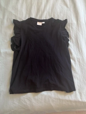 Svart ärmlös topp med volang - Svart ärmlös topp med volangdetaljer på axlarna. Rund halsringning och rak passform. Perfekt för en stilren och trendig look. Mjuk och bekväm känsla.