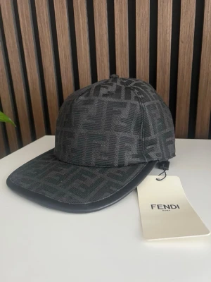 Svart keps med Fendi-logga - Snygg svart keps från Fendi med klassiskt FF-mönster över hela kepsen. Kepsen har böjd skärm och är tillverkad i ett strukturerat tyg som ger en exklusiv känsla. Perfekt för dig som vill ha en trendig accessoar med lyxig touch.