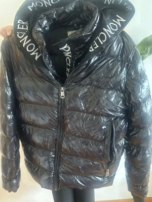 Svart pufferjacka från Moncler - Snygg svart pufferjacka från Moncler med glansig finish och stor huva med Moncler-logga i vitt. Jackan har dragkedja framtill, två sidofickor med dragkedja och quiltad design. Perfekt för kalla dagar när du vill hålla stilen. Finns fåtal defekter som vid armen och ett hål i ena fickan.