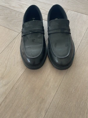 Svarta loafers från Next - Snygga svarta loafers från Next med klassisk penny strap och rundad tå. Skorna är tillverkade i slätt syntetmaterial och har en låg klack samt diskret sömdetalj runt sulan. Perfekta för dig som vill ha en stilren och tidlös look.