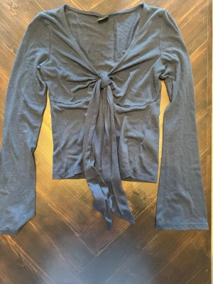 Blå omlottopp med knyt - Trendig blå omlottopp med långa ärmar och knyt framtill. Toppen har en djup v-ringning och är tillverkad i ett mjukt, stretchigt material som sitter snyggt på kroppen. Perfekt för att skapa en cool och avslappnad look.