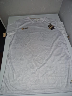 Vit AIK t-shirt med guld och svart - Snygg vit t-shirt från AIK med diskret stadsmotiv i tyget. Guld- och svartfärgade detaljer på ärmarna, AIK-emblem på bröstet och Stockholm-märke nedtill. Perfekt för dig som vill visa ditt AIK-stöd med stil.