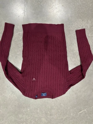 Gant tröja - Vinröd kabelstickad tröja från Gant med rund halsringning och diskret logga broderad på bröstet. Tröjan har långa ärmar och ribbade muddar vid ärmslut och nederkant. Klassisk och tidlös design i mjukt material.