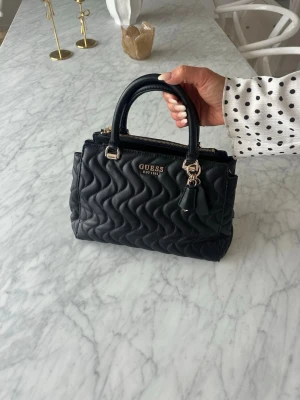 Svart quiltad handväska från Guess - Säljer en svart quiltad handväska från Guess med guldfärgade detaljer och dubbla handtag. Väskan har vågigt mönster, dragkedja upptill och en dekorativ nyckelring. Perfekt för att bära dina viktigaste saker.