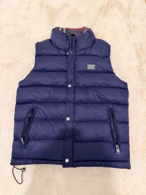 Mörkblå dunväst från Burberry - Snygg mörkblå dunväst från Burberry med hög krage och två dragkedjeförsedda fickor. Västen har quiltad design och ett Burberry London England-märke på bröstet. Perfekt för lager på lager och håller dig varm under kyliga dagar.
