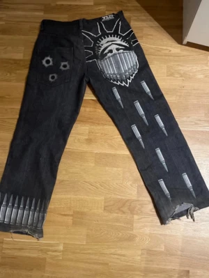 Yld jeans  - Unika svarta jeans med handmålat motiv av öga, taggar och patroner. Coola detaljer som kulhål på bakfickan och slitna benslut. Rak passform och riktigt statement-plagg för dig som vill sticka ut.