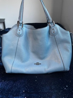 Blå läderhandväska från Coach - Snygg ljusblå handväska från Coach i äkta läder med dubbla handtag och silverfärgade detaljer. Väskan har en klassisk och rymlig design med Coach-loggan framtill. Perfekt för dig som vill ha en stilren och trendig accessoar.