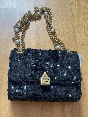 Svart paljettväska från Guess - Supersnygg svart handväska från Guess täckt med glittriga paljetter. Väskan har en guldfärgad kedja med stora pärldetaljer och ett dekorativt lås framtill. Perfekt för dig som vill sticka ut och addera lite extra bling till din outfit.