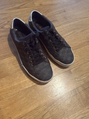 Svarta sneakers från Michael Kors - Snygga svarta sneakers från Michael Kors med diskret mönster och vita sulor. Skorna har klassisk låg modell, svarta snören och silverdetaljer vid hälen. Perfekta för dig som vill ha en stilren och trendig look.