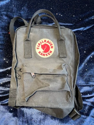 Svart Kånken ryggsäck från Fjällräven - Klassisk svart Kånken ryggsäck från Fjällräven med rund röd och vit logotyp framtill. Ryggsäcken har ett stort huvudfack, en mindre ficka med dragkedja på framsidan och justerbara axelband. Materialet är slitstark syntetväv.