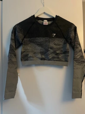 Gymshark crop top och långärmad tröja - Två snygga träningsplagg från Gymshark: en svart crop top med stort vitt Gymshark-tryck framtill och en grå/svart långärmad crop tröja med mönster och logga på bröstet. Perfekt för gymmet eller sportaktiviteter.