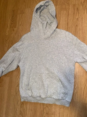 Ljusgrå hoodie med huva - En basic ljusgrå hoodie med stor huva och magficka. Tillverkad i mjukt sweatshirtmaterial, perfekt för chill dagar. Klassisk passform med ribbade muddar vid ärmslut och nederkant. Inga synliga loggor eller tryck.