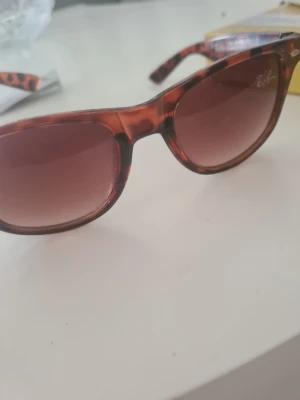 Ray-Ban leopardmönstrade solglasögon - Snygga solglasögon från Ray-Ban med leopardmönstrade bågar i brun och svart. Glasen är mörka och har en rundad form som ger en cool look. Perfekt för att höja stilen under soliga dagar.