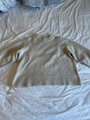 Beige stickad tröja med ribbad kant - En enkel och stilren beige stickad tröja med rund hals och ribbad kant vid nederdelen och ärmslut. Tröjan har lång ärm och är gjord i ett mjukt material som passar perfekt till hösten och vintern.