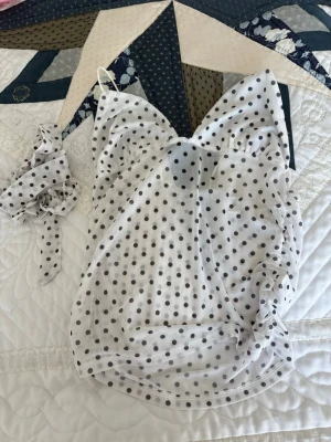 Vit prickig linnetopp med rosett - Söt vit linnetopp med svarta prickar och tunna axelband. Toppen har en matchande rosett som kan användas i håret eller som accessoar. Materialet känns lätt och luftigt, perfekt för varma dagar. Trendig och lekfull design som sticker ut.