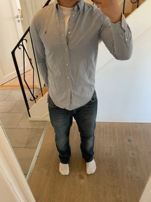 Ljusblå skjorta från Polo Ralph Lauren - Säljer en klassisk ljusblå skjorta från Polo Ralph Lauren med knappar framtill och långärmad design. Skjortan har en broderad logotyp på bröstet och är tillverkad i bomull. Perfekt för dig som gillar stilrena plagg med snygga detaljer.