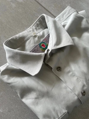 Ljusgrå skjorta från Fjällräven vintage - Stilren ljusgrå skjorta från Fjällräven med klassisk krage och två bröstfickor med knappar. Skjortan har långa ärmar och är tillverkad i ett slitstarkt material, perfekt för dig som gillar outdoor eller vill ha en clean look. Den har också två små skador vid hålen, kan skicka bild i dm