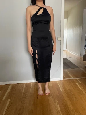 Svart satin klänning med asymmetrisk remdetalj och slits - Snygg svart klänning i satin med unik cut-out framtill och smala axelband. Klänningen har en slits med snörningsdetalj på framsidan och en figurnära passform. En dold dragkedja i sidan.