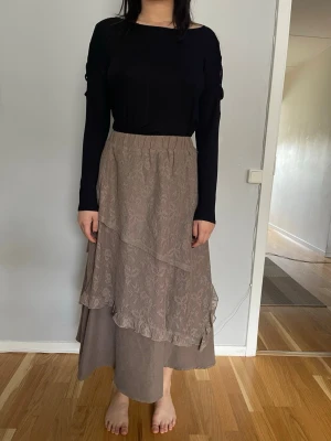 Beige långkjol med volang - Säljer en beige långkjol med dubbla lager och volangdetaljer. Kjolen har ett subtilt blommigt mönster på det övre lagret och en elastisk midja som gör den bekväm att bära. Köpt i Sydkorea och har aldrig använtNy med prislapp..
