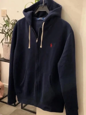 Mörkblå hoodie från Ralph Lauren - Säljer en snygg mörkblå hoodie från Ralph Lauren med dragkedja och vit snörning i huvan. Den har en röd broderad logga på bröstet och praktiska fickor framtill. Perfekt för en avslappnad stil. Kan passa S också! 