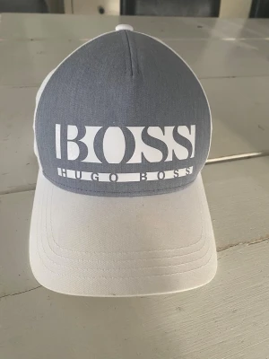 Gråvit keps från Hugo Boss - Snygg keps från Hugo Boss med grå frontpanel och vit skärm samt baksida. Stor vit BOSS-logga framtill och justerbar rem baktill. Materialet är bomull och kepsen har en klassisk form med böjd skärm.