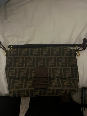 brun handväska - Snygg brun handväska  i mörkbrunt och beige. Väskan har ett lock med brunt spänne och axelrem i läder. Guldiga metalldetaljer och rymligt innerfack. Perfekt för dig som gillar lyxiga accessoarer och tidlös design.