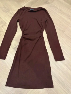 Brun långärmad klänning med rynk - Säljer en brun långärmad klänning från NA-KD med snygg rynkdetalj i midjan som ger en smickrande siluett. Klänningen är i mjukt, stretchigt material och har en enkel, stilren design som passar perfekt till olika tillfällen.