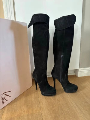 Svarta overknee stövlar från K.Cobler - Säljer ett par svarta overknee stövlar från K.Cobler med hög klack och platåsula. Stövlarna är tillverkade i mocka och har en snygg vikkant upptill samt dragkedja på sidan. Perfekt för dig som vill ha en statement-look. Insidan an fodret har luckrats upp men påverkar inte utseendet. 