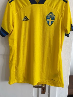 Svensk landslagströja Adidas gul - Gul fotbollströja från Adidas med blå detaljer och Sveriges landslagsemblem på bröstet. Tröjan har korta ärmar med blå ränder och en mörkblå krage. Materialet är lätt och andas, perfekt för sport och träning.  (Authentic)