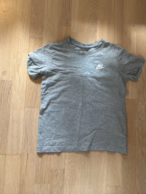 Grå Nike t-shirt 140 - Säljer en enkel och stilren grå t-shirt från Nike med klassisk vit logga på bröstet. T-shirten har rund hals och korta ärmar, perfekt för dig som gillar sportiga och avslappnade looks. Materialet är mjukt och känns skönt mot huden.