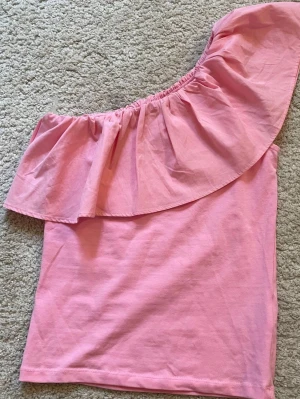 Rosa oneshoulder topp med volang - Säljer en snygg rosa oneshoulder topp med stor volang längs halsen. Perfekt för dig som gillar att sticka ut och vill ha något trendigt. Toppens material är mjukt och lätt bomull, vilket gör den bekväm att bära. Stolek 170💕