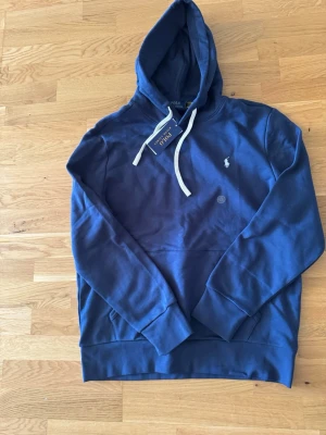 Blå hoodie från Polo Ralph Lauren - Snygg blå hoodie från Polo Ralph Lauren med vit dragsko och klassisk logga broderad på bröstet. Hoodien har en stor huva, ribbade muddar och en praktisk magficka. Perfekt för dig som gillar stilren design och mjukt material.