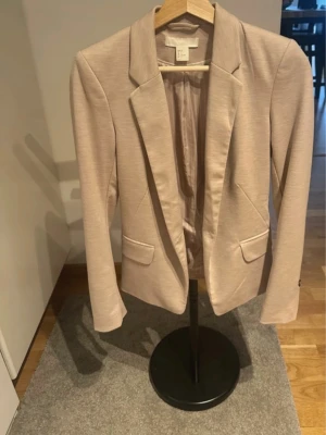 Beige kavaj från H&M med fickor - Snygg beige kavaj från H&M med klassisk passform och två framfickor med lock. Kavajen har smala slag och är helfodrad. Perfekt för dig som vill ha en stilren look. Materialet är mjukt och lätt, vilket gör den bekväm att bära.