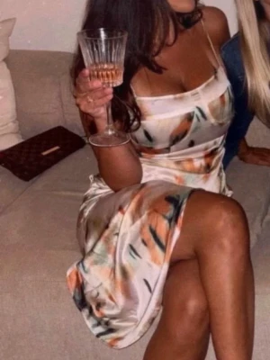 Mönstrad satin klänning med smala band - Säljer en elegant klänning i satin med smala axelband och abstrakt mönster i beige, svart och orange toner. Klänningen har en mjuk, glansig finish och är perfekt för dig som vill ha en stilren look med lite extra färg.