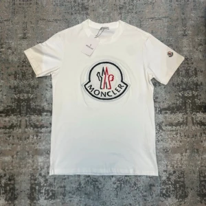 Vit t-shirt med stor logga - Snygg vit t-shirt med stor broderad logga i svart och rött på bröstet. Klassisk passform och rund halsringning. Tillverkad i mjuk bomull och har en liten patch på ärmen. Perfekt för dig som gillar stilrena märkesplagg.