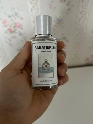 Sabatier 3.0 Extrait de Parfum - Säljer en stilren parfymflaska från One Bold Chemist, modellen Sabatier 3.0. Flaskan är genomskinlig med en silvrig metallkork och har en modern etikett med grafiskt tryck. Innehållet är ljusbrunt och flaskan har en klassisk rund form.