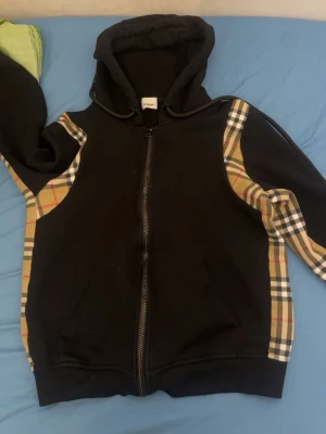 Svart hoodie med rutiga detaljer - Snygg svart hoodie med dragkedja och klassiska beige-rutiga paneler längs ärmarna och axlarna. Hoodien har en stor huva och två fickor framtill. Perfekt för dig som gillar streetwear och vill sticka ut lite extra.