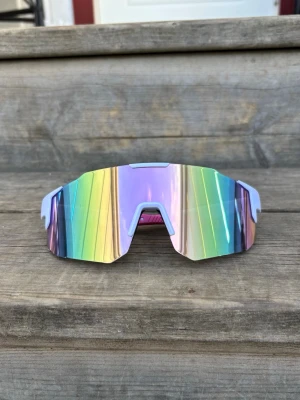 Pastellrosa Sportsolglasögon med vit ram - Säljer ett par riktigt coola solglasögon med stora, spegelblanka linser i regnbågsfärger och randigt mönster. De har en sportig och futuristisk design med ljusblå detaljer runt linserna. Perfekta för dig som vill sticka ut och ha en unik look.