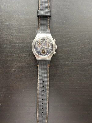 Swatch svart kronograf herrklocka - Snygg Swatch kronograf med svart urtavla och orange detaljer. Boetten är i silverfärgad metall och armbandet är svart med orange sömmar, tillverkat i syntetmaterial. Klockan har flera små urtavlor och klassisk rund form.