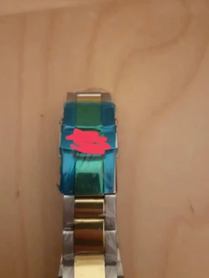 Stilren klocka med färgad boett - Snygg klocka med metallarmband i silver och guldton. Urtavlan är svart med färgglad ring i rött och blått, samt tydliga siffror. Klockan har en klassisk rund form och metallspänne. Perfekt accessoar för dig som vill sticka ut lite extra.