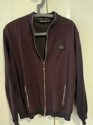 Mörklila bomberjacka Emporio Armani - Snygg mörklila bomberjacka från Emporio Armani med silverfärgade dragkedjor och ribbade muddar. Jackan har en klassisk bombermodell med rund halsringning och två fickor framtill. Märkets logga sitter diskret på bröstet.