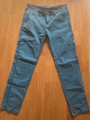 Blå chinos med knappdetaljer - Snygga blå chinos med raka ben och klassiska fickor fram och bak. Byxorna har knappdetaljer vid fickorna och stängs med knapp och dragkedja. Perfekta för dig som vill ha en stilren och bekväm look. Materialet är mjukt och lätt bomull.