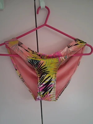 Tropiska bikiniunderdel i rosa och gul - Säljer en tropisk bikiniunderdel med färgglatt palmbladsmönster i rosa, gul och svart. Modellen har hög skärning och är tillverkad i mjukt syntetmaterial som passar perfekt för strandhäng och soliga dagar. Aldrig använd