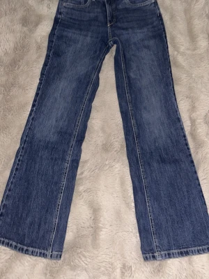 Blå bootcut jeans med kontrastsömmar - Säljer ett par klassiska blå bootcut jeans med kontrastsömmar och femficksdesign. Jeansen har en normal midja och dragkedja med knapp framtill. Materialet är denim och passformen är något utsvängd nedtill.